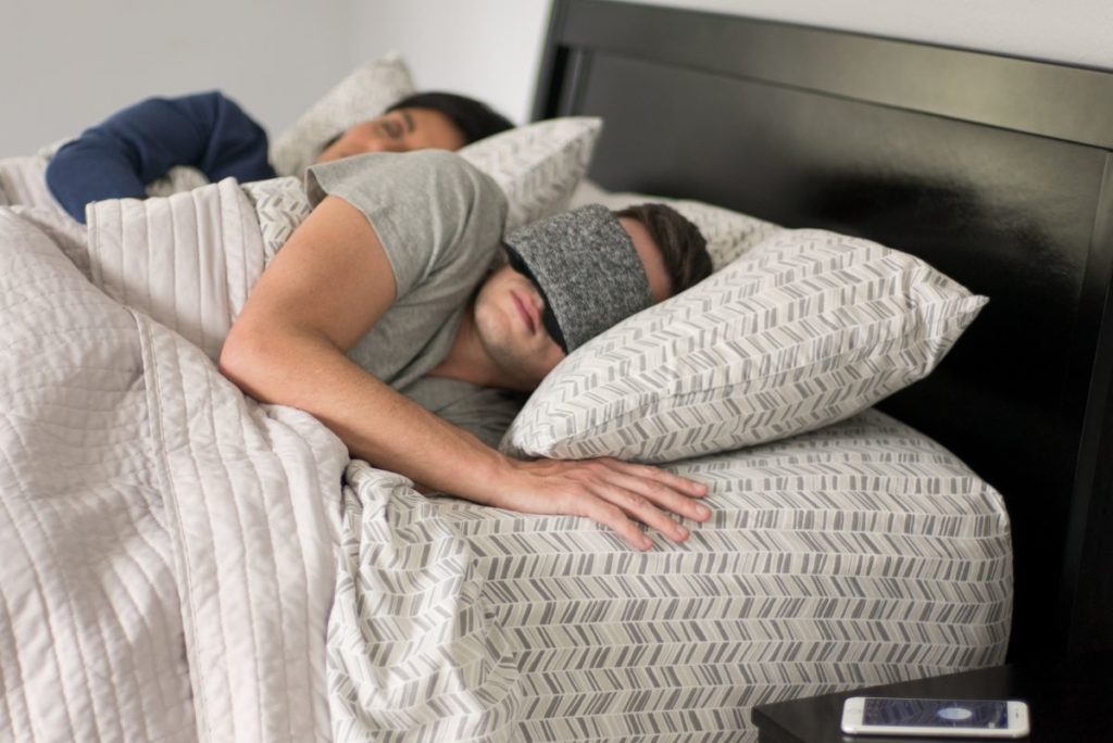Zwei Menschen liegen schlafend im Bett. Eine Person trägt die Schlafmaske von Hupnos. Auf dem Nachtisch liegt ein Handy.