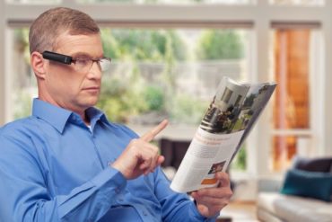 Ein älterer Mann sitzt im Stuhl. Er trägt eine Brille, an der die OrCam MyEye befestigt ist und zeigt auf eine Zeitung, die er in der Hand hält.