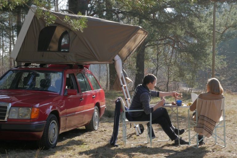 Das Dachzelt steht auf einem Auto im Wald. Davor sitzen zwei junge Leute und trinken etwas.