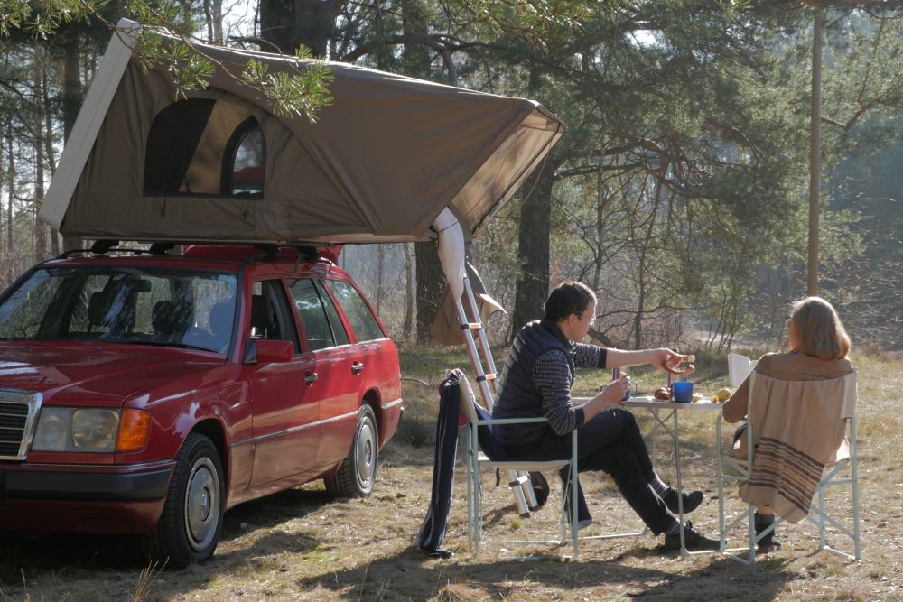 Das Dachzelt steht auf einem Auto im Wald. Davor sitzen zwei junge Leute und trinken etwas.